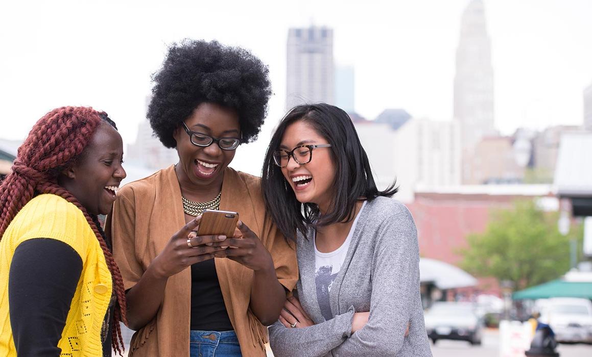 三名有色人种女性学生站在一起对着电话微笑，背景是堪萨斯城的天际线